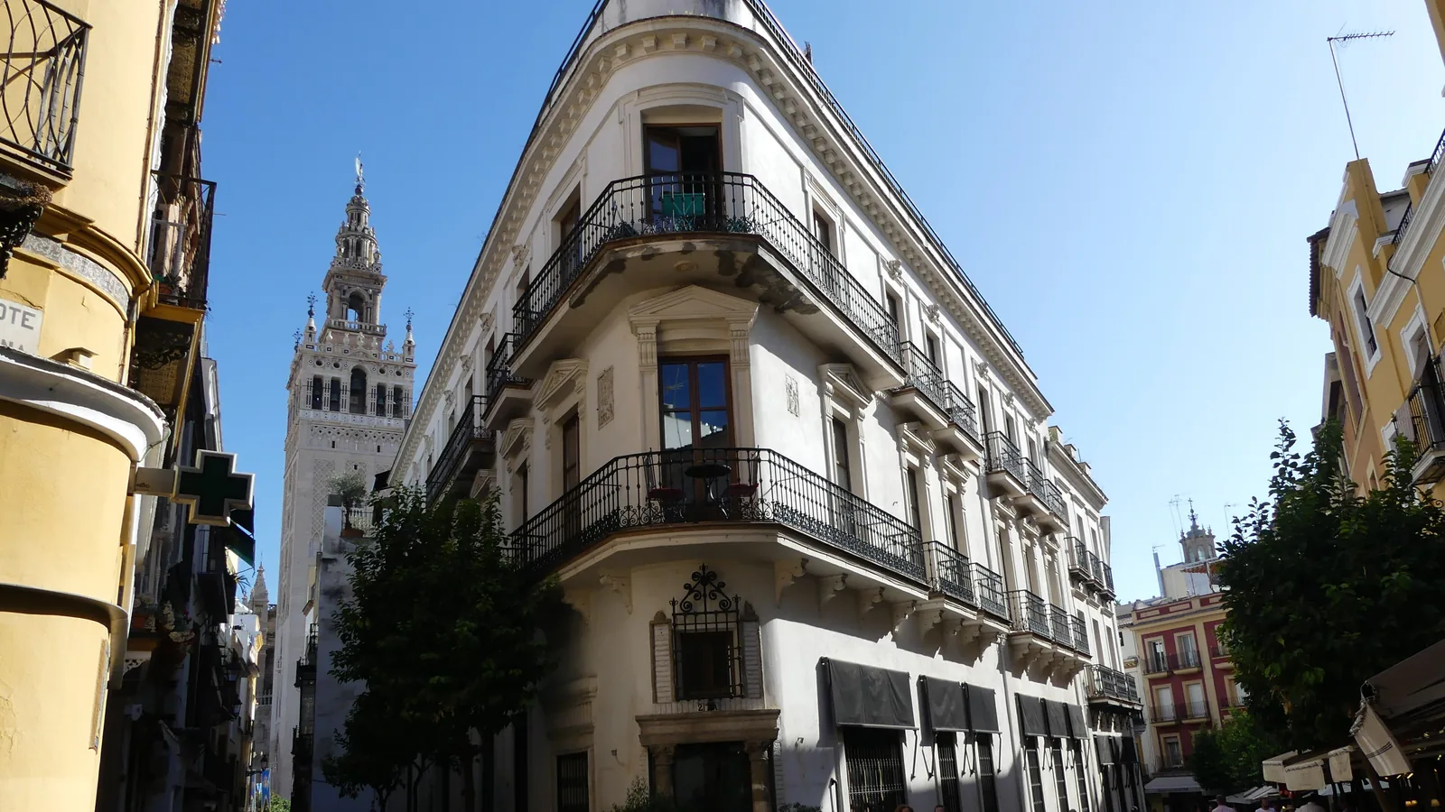 A street corner in central Seville with a balconied corner building and the Giralda tower rising behind it under a blue sky